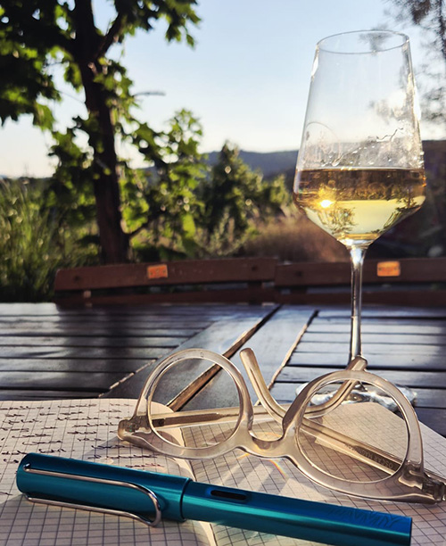 Glas Wein auf einem Tisch mit einem geöffneten Schreibheft, auf dem ein Schreibstift liegt. Im Hintergrund ein Weingarten in der Abendsonne